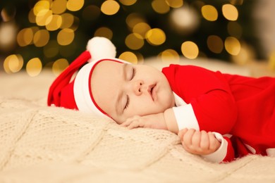 Photo of Cute baby in Christmas costume sleeping on knitted blanket against blurred festive lights. Winter holiday