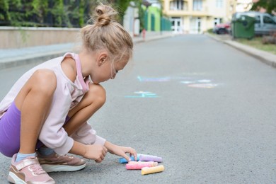 Photo of Little child with chalk pieces sitting on asphalt outdoors, space for text
