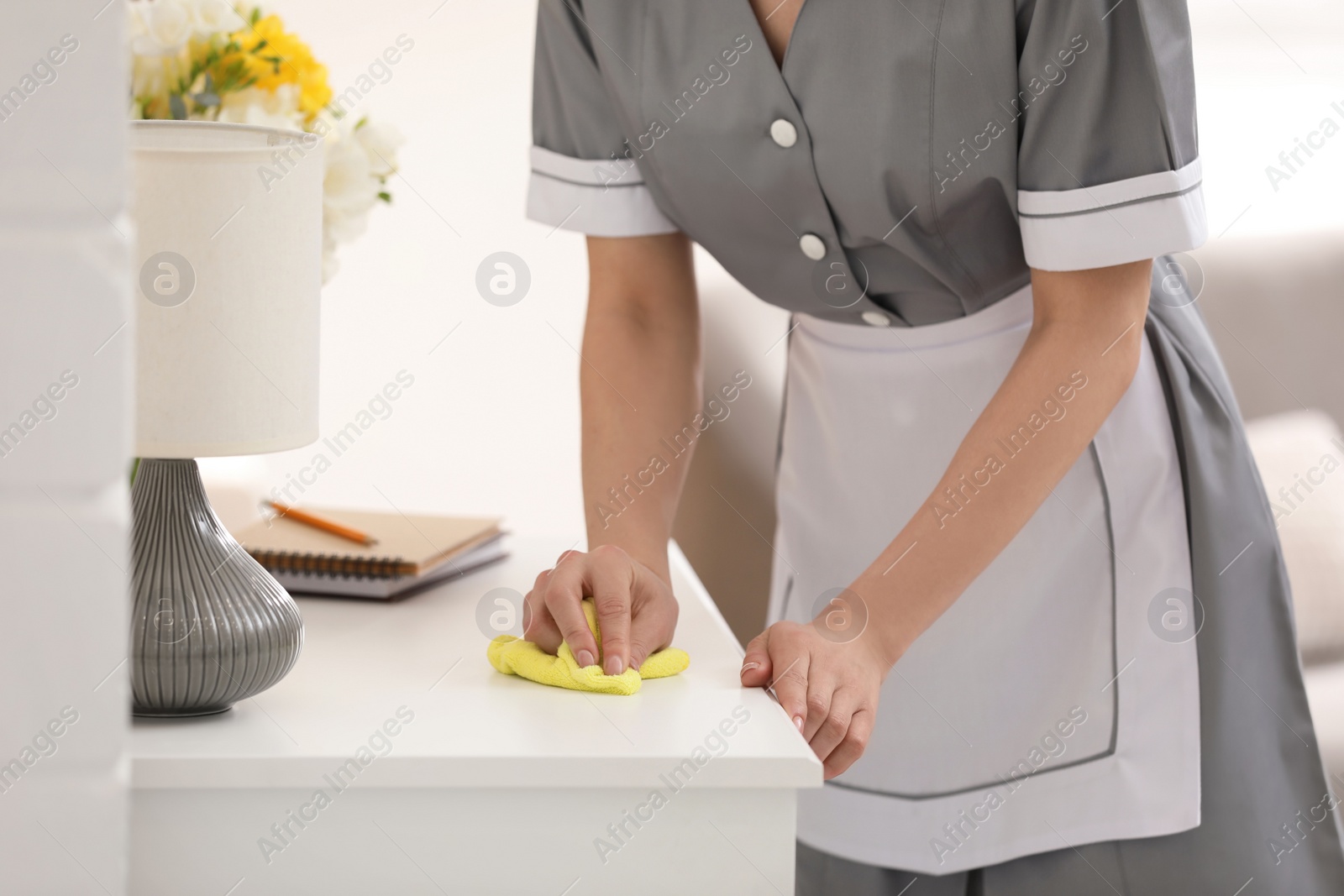 Photo of Young chambermaid wiping dust from furniture in hotel room, closeup