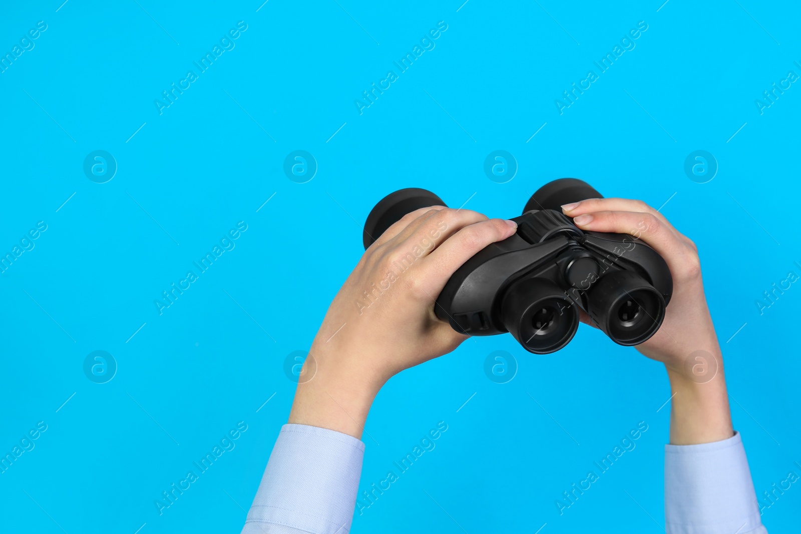 Photo of Woman holding modern binoculars on light blue background, closeup. Space for text