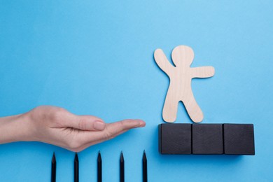 Photo of Woman holding hand to help human figure avoid trap with pencils as spikes on light blue background, closeup