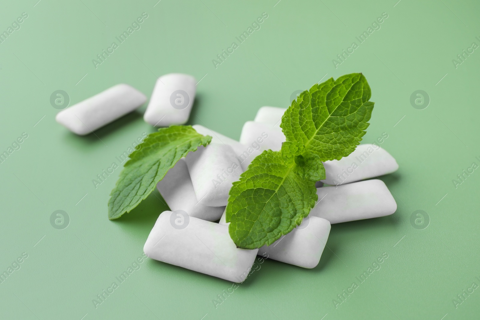 Photo of Tasty white chewing gums and mint leaves on light green background, closeup