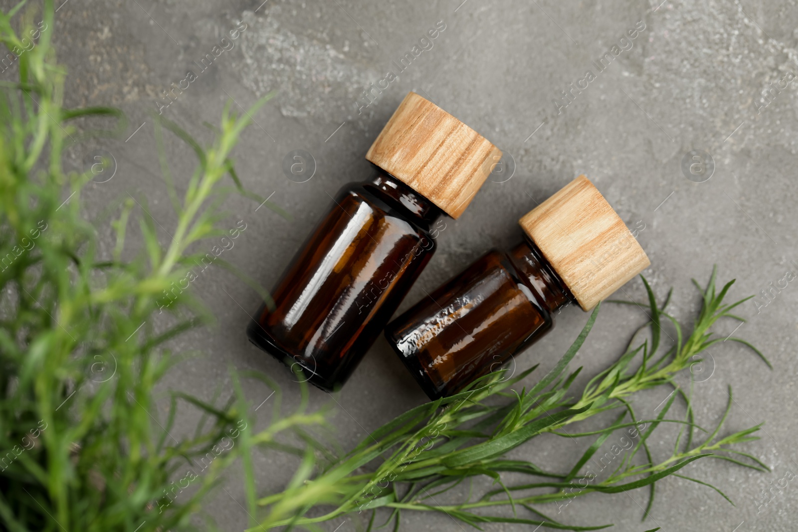 Photo of Bottles of essential oil and fresh tarragon leaves on grey table, flat lay