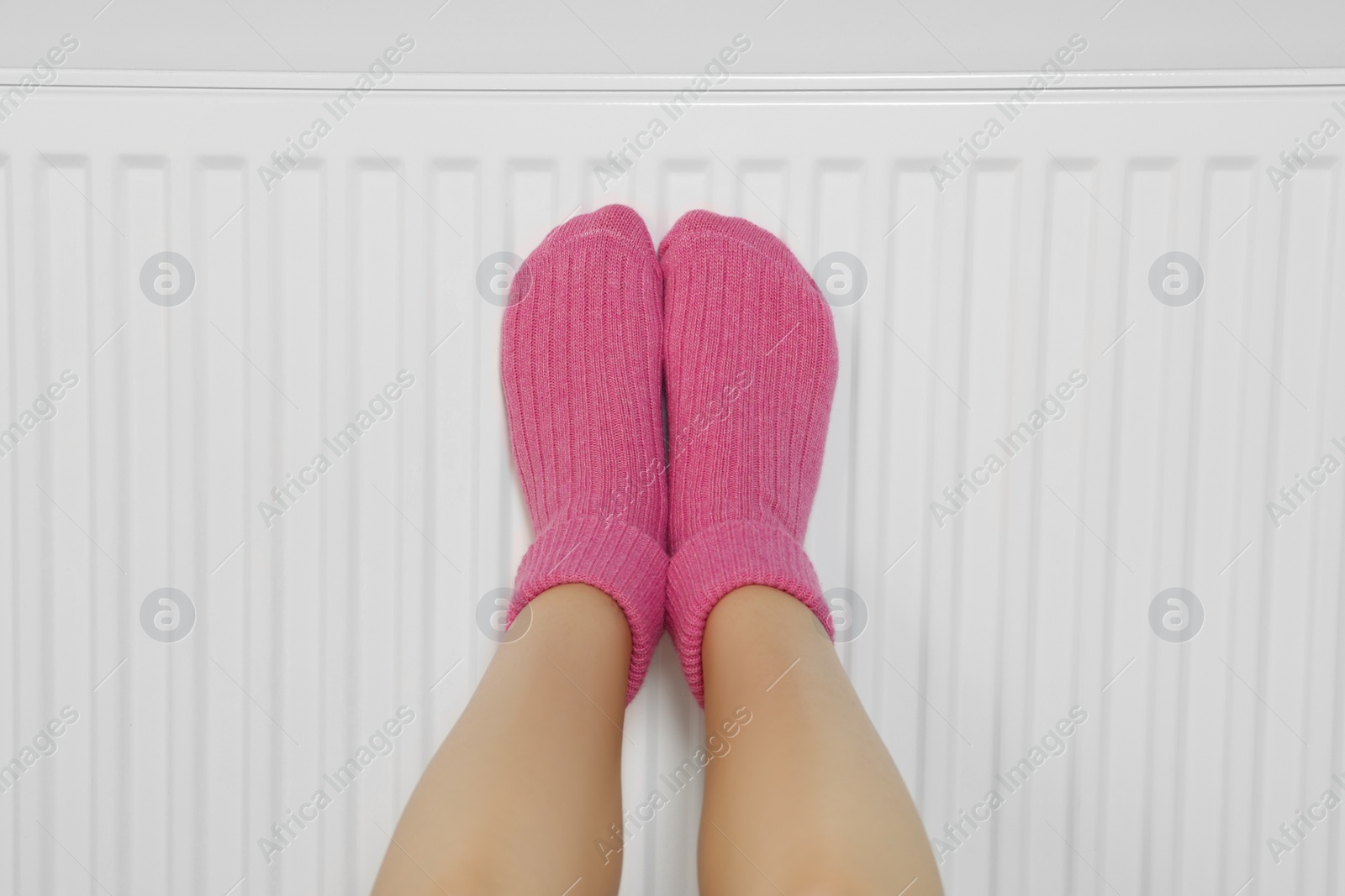 Photo of Woman warming legs on heating radiator near white wall, closeup