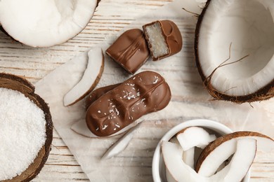 Delicious milk chocolate candy bars with coconut filling on white wooden table, flat lay