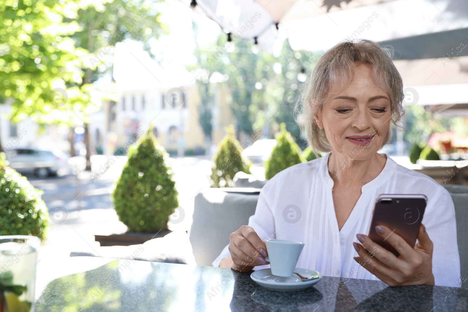 Photo of Mature woman enjoying tasty coffee while using mobile phone in cafe on sunny day. Space for text