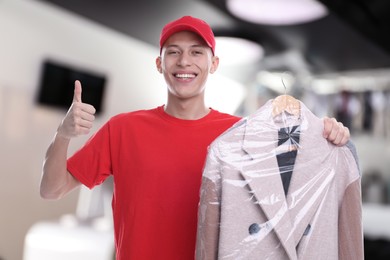 Happy courier holding coat in plastic bag and showing thumbs up in dry-cleaning