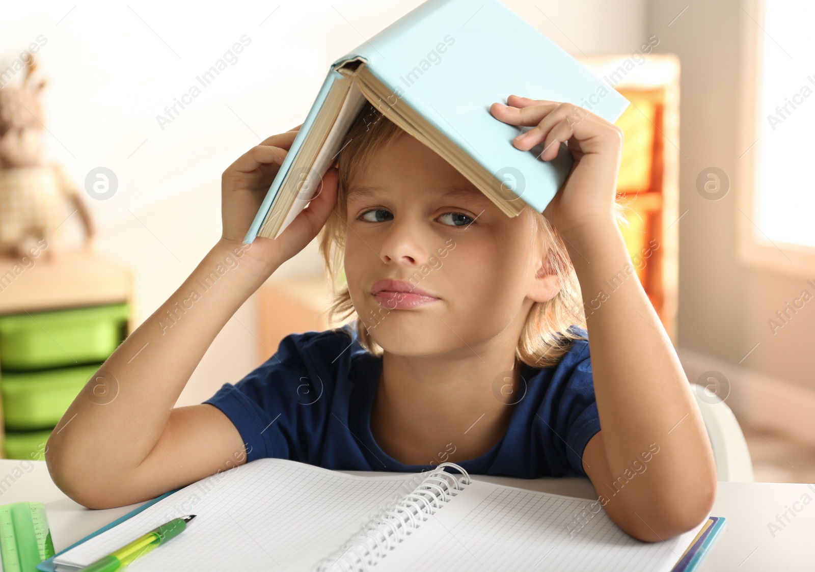 Photo of Bored little boy with book on his head doing homework at table indoors