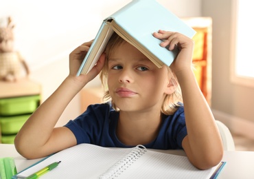 Photo of Bored little boy with book on his head doing homework at table indoors