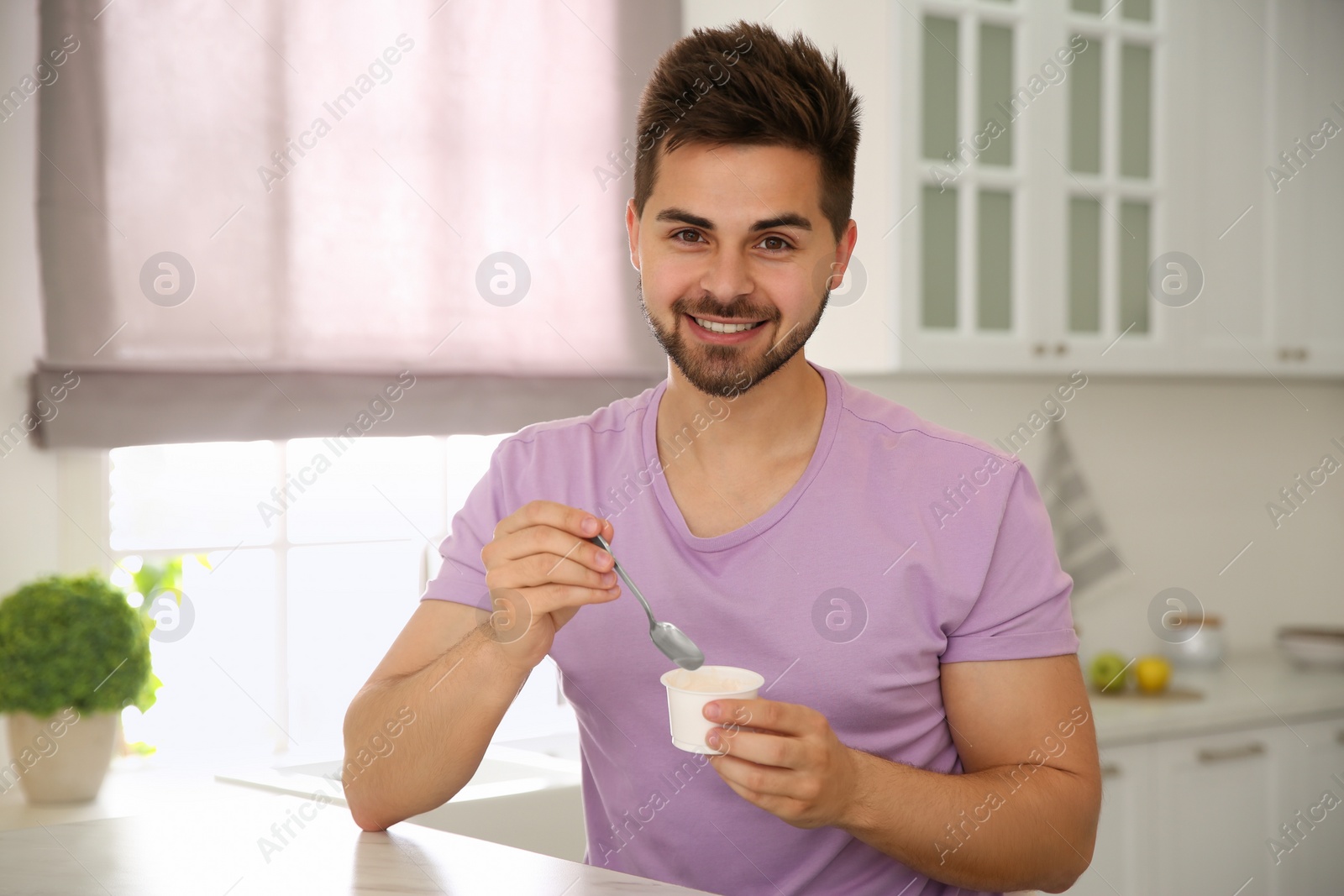 Photo of Happy young man with tasty yogurt at table in kitchen