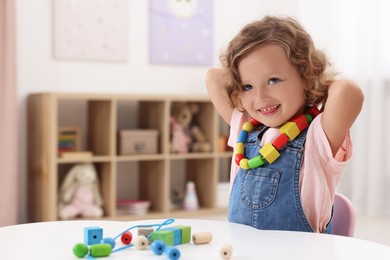 Photo of Motor skills development. Little girl playing with wooden pieces and string for threading activity at table indoors