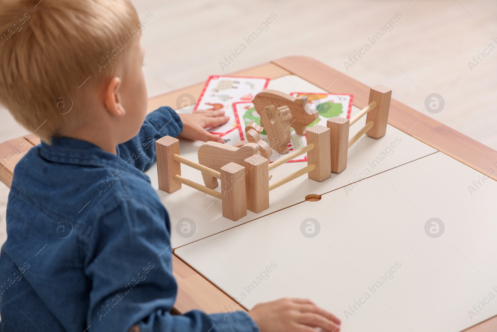 Photo of Little boy playing with set of wooden animals and fence at table indoors. Child's toy