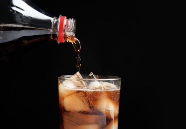 Photo of Pouring refreshing cola from bottle into glass with ice cubes on black background, closeup