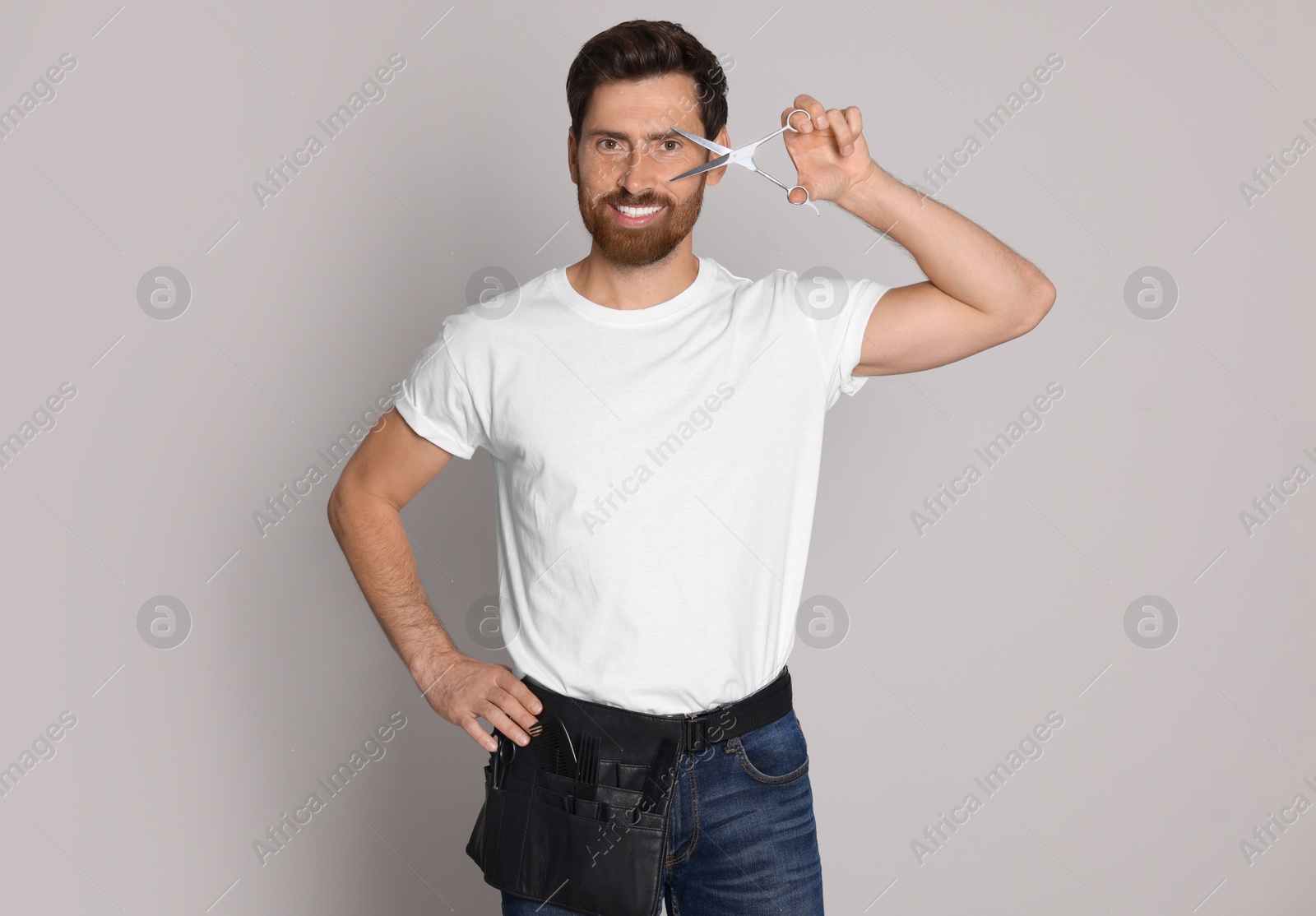 Photo of Smiling hairdresser with tool bag holding scissors on light grey background