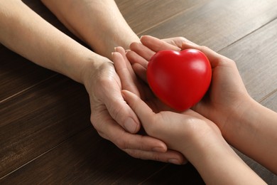Photo of Young and elderly women holding red heart at wooden table, closeup