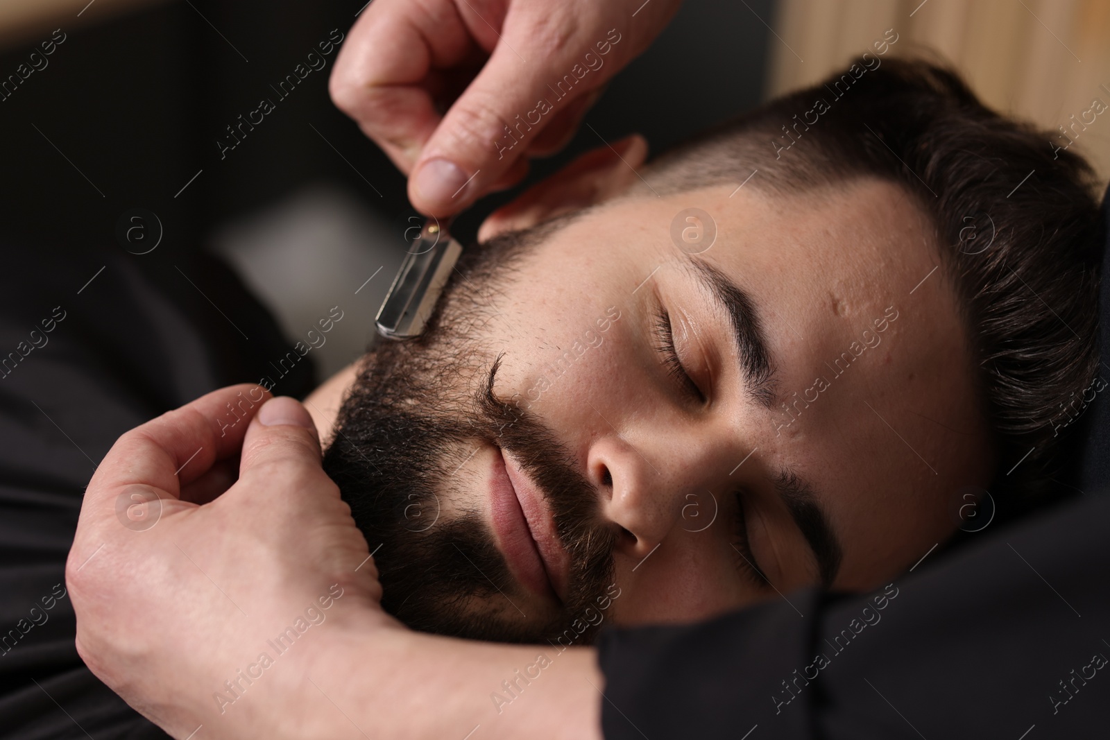 Photo of Professional barber shaving client's beard with blade in barbershop, closeup