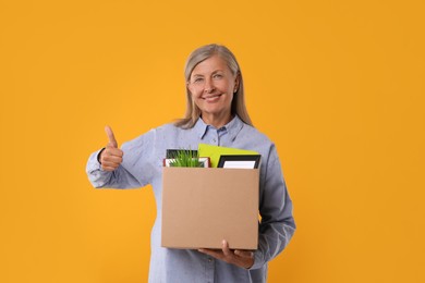 Photo of Happy unemployed senior woman with box of personal office belongings showing thumb up on orange background
