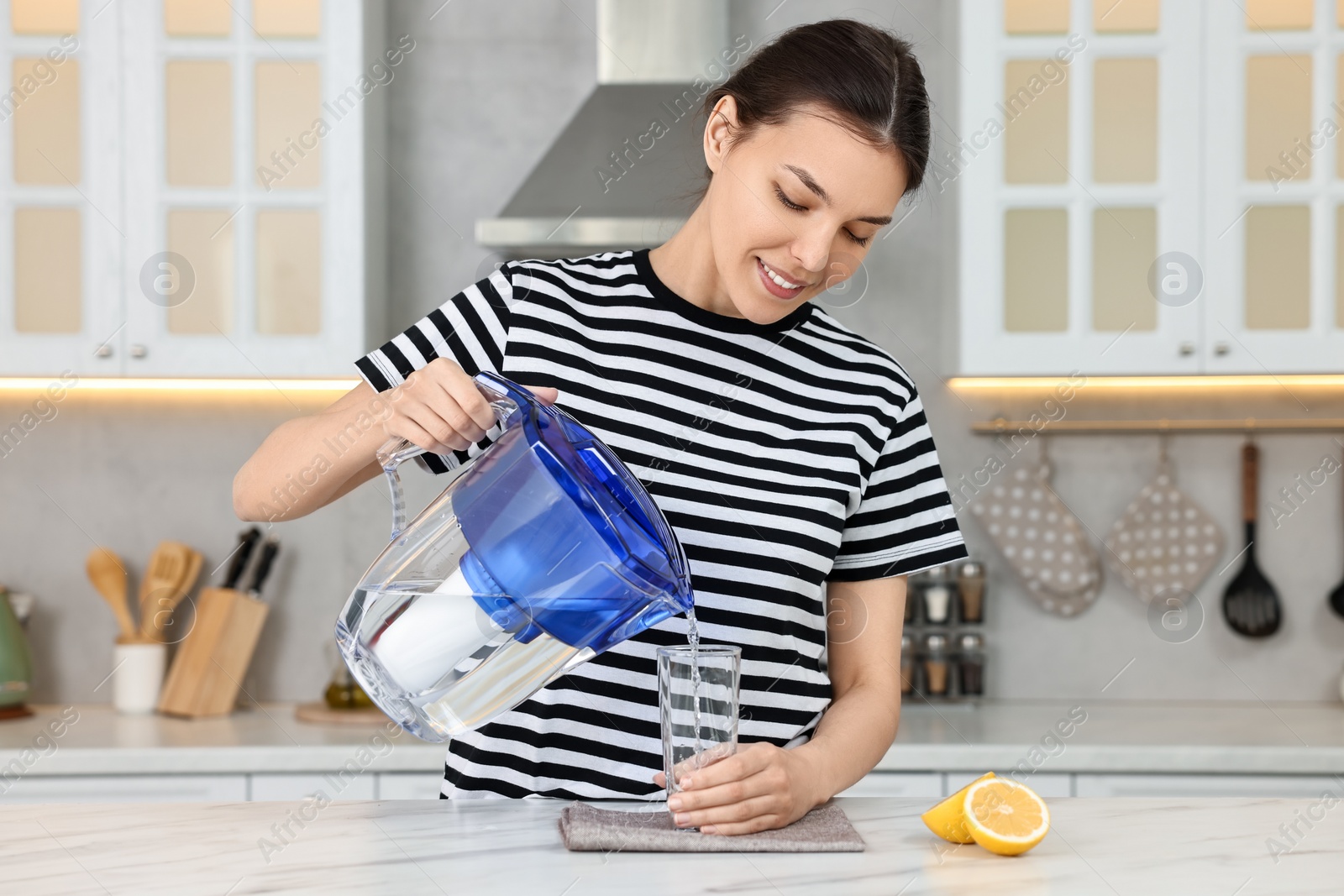 Photo of Woman pouring water from filter jug into glass in kitchen