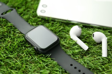 Photo of Stylish smart watch, phone and earphones on green grass, closeup