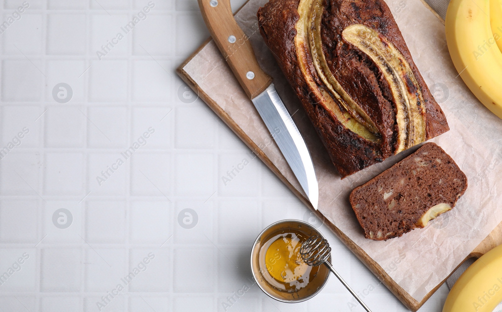 Photo of Delicious banana bread, honey and knife on white tiled table, flat lay. Space for text