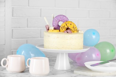 Delicious cake decorated with sweets, tableware and balloons on white marble table