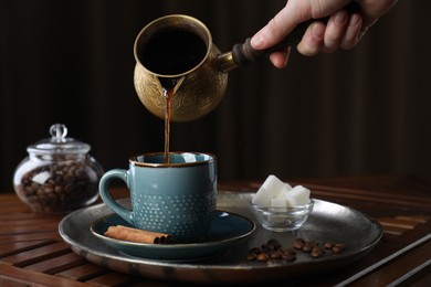 Turkish coffee. Woman pouring brewed beverage from cezve into cup at wooden table, closeup