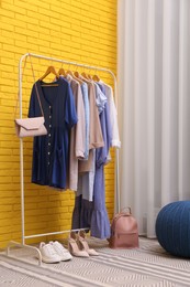 Rack with different stylish clothes and shoes near yellow brick wall in room