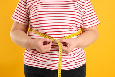 Overweight woman measuring waist with tape on orange background, closeup