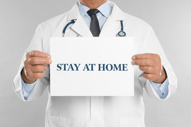 Image of Doctor holding sign with text STAY AT HOME on light grey background, closeup. Quarantine during coronavirus outbreak