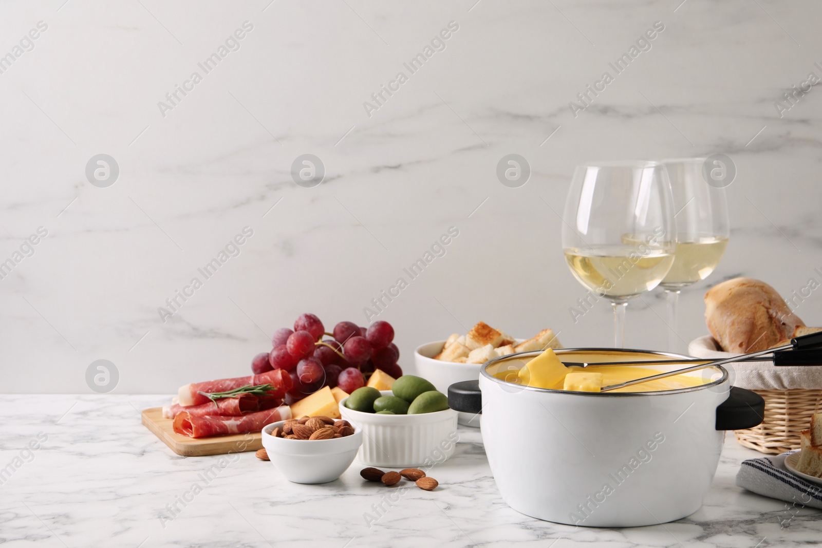 Photo of Pot of tasty cheese fondue with snacks and wine on white marble table, space for text