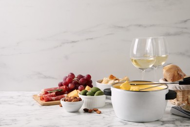 Pot of tasty cheese fondue with snacks and wine on white marble table, space for text