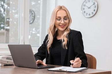 Happy secretary taking notes while working with laptop at table in office