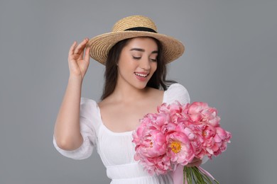 Photo of Beautiful young woman in straw hat with bouquet of pink peonies against grey background