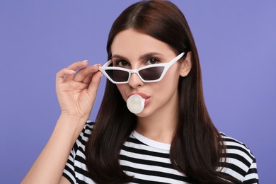 Photo of Beautiful woman in sunglasses blowing bubble gum on light purple background