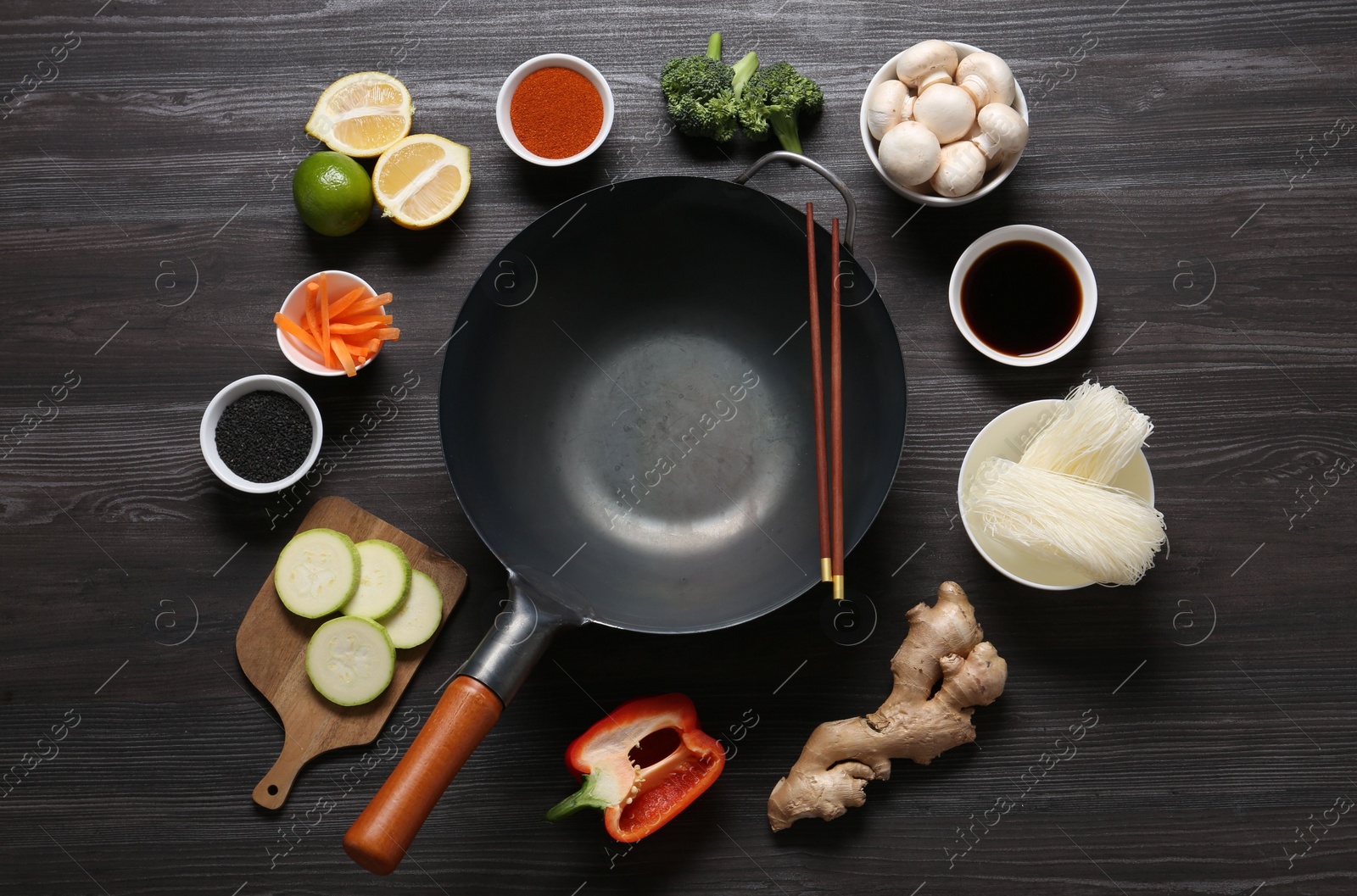 Photo of Empty iron wok and chopsticks surrounded by ingredients on dark grey wooden table, flat lay