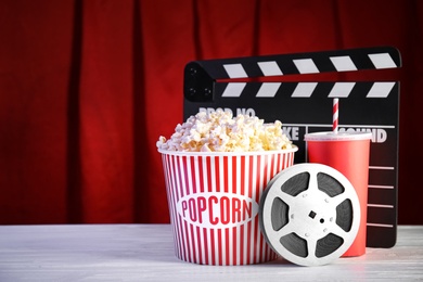 Clapperboard, reel and popcorn bucket on table against stage curtain, space for text. Watching cinema