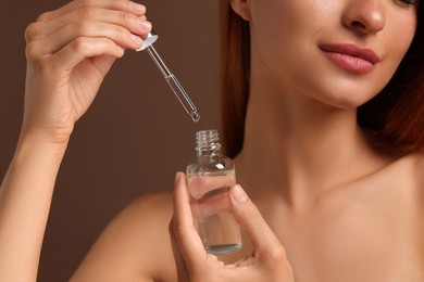 Beautiful young woman with bottle of cosmetic serum on brown background, closeup