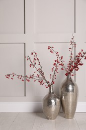 Photo of Hawthorn branches with red berries in vases near light  wall indoors