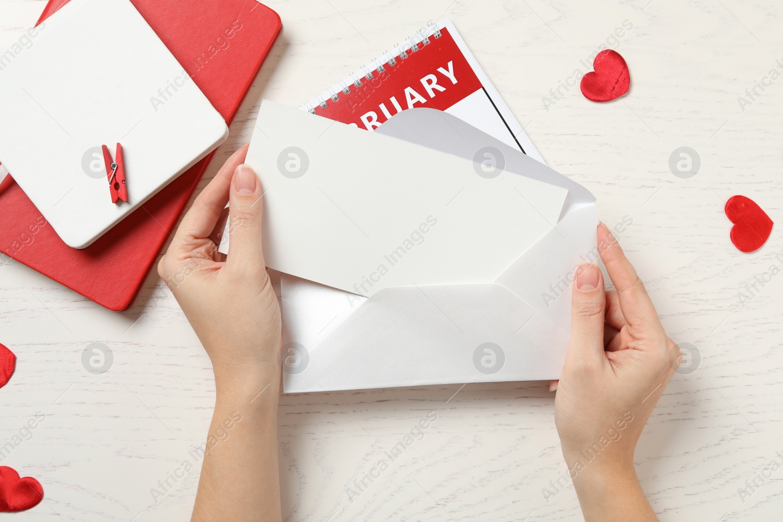 Photo of Woman putting blank greeting card into envelope above white wooden table, top view. Valentine's day celebration