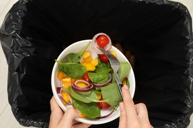 Woman throwing vegetable salad into bin indoors, top view