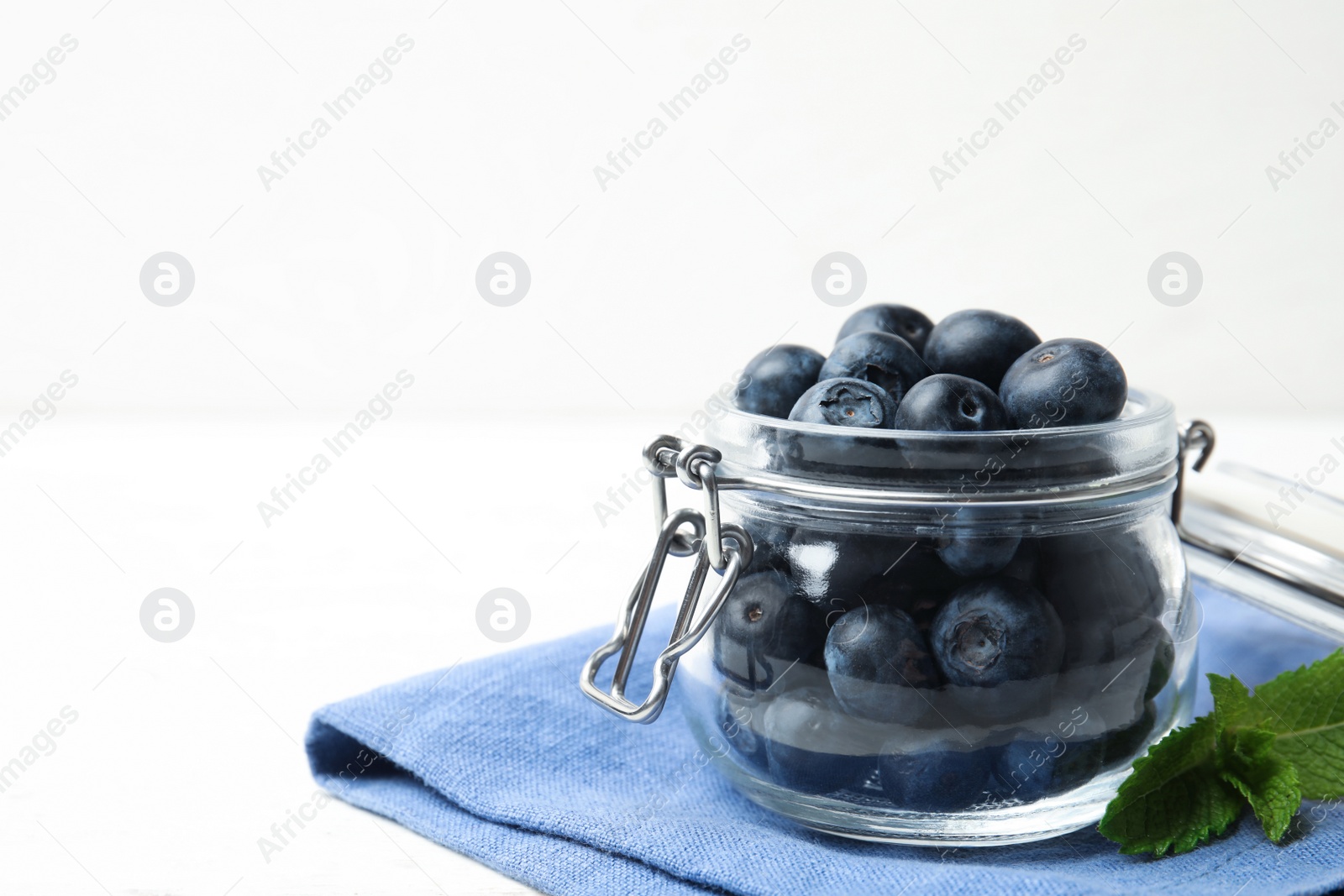 Photo of Glass jar of tasty fresh blueberries, mint leaves and fabric on white wooden table, space for text