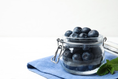 Photo of Glass jar of tasty fresh blueberries, mint leaves and fabric on white wooden table, space for text