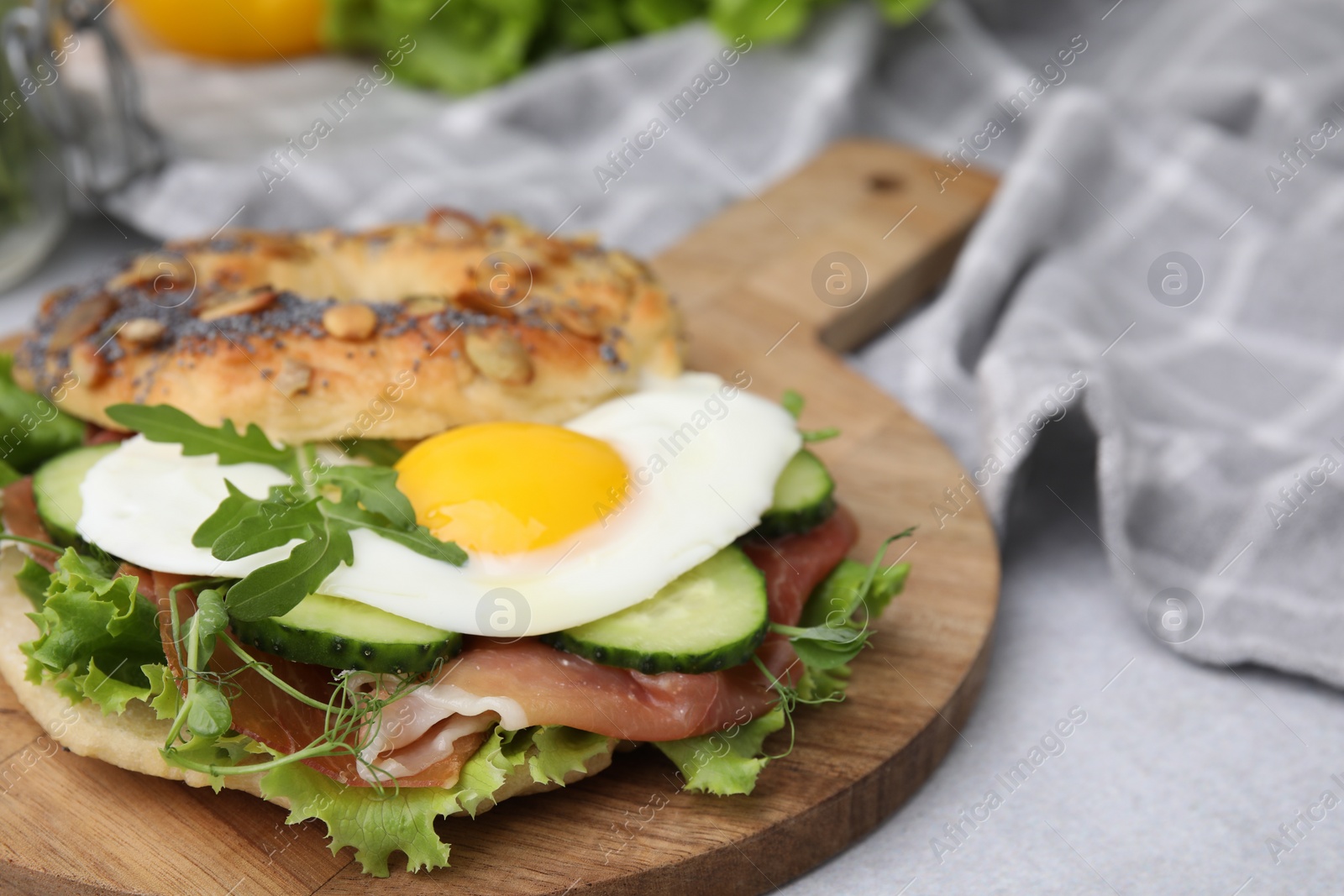 Photo of Tasty bagel with cured ham, egg, cucumber and salad mix on light table, closeup. Space for text