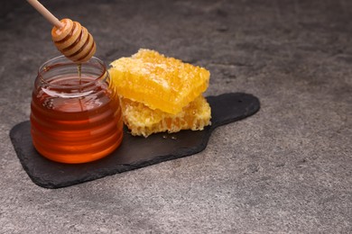 Pouring sweet golden honey from dipper into jar and pieces of honeycomb on grey textured table, space for text