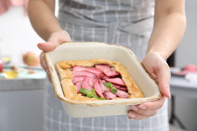 Photo of Woman holding baking dish with delicious apple galette indoors, closeup