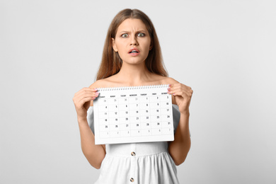 Emotional young woman holding calendar with marked menstrual cycle days on light background