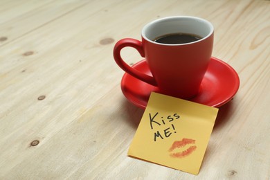 Photo of Sticky note with phrase Kiss Me, lipstick mark and cup of coffee on wooden table, space for text