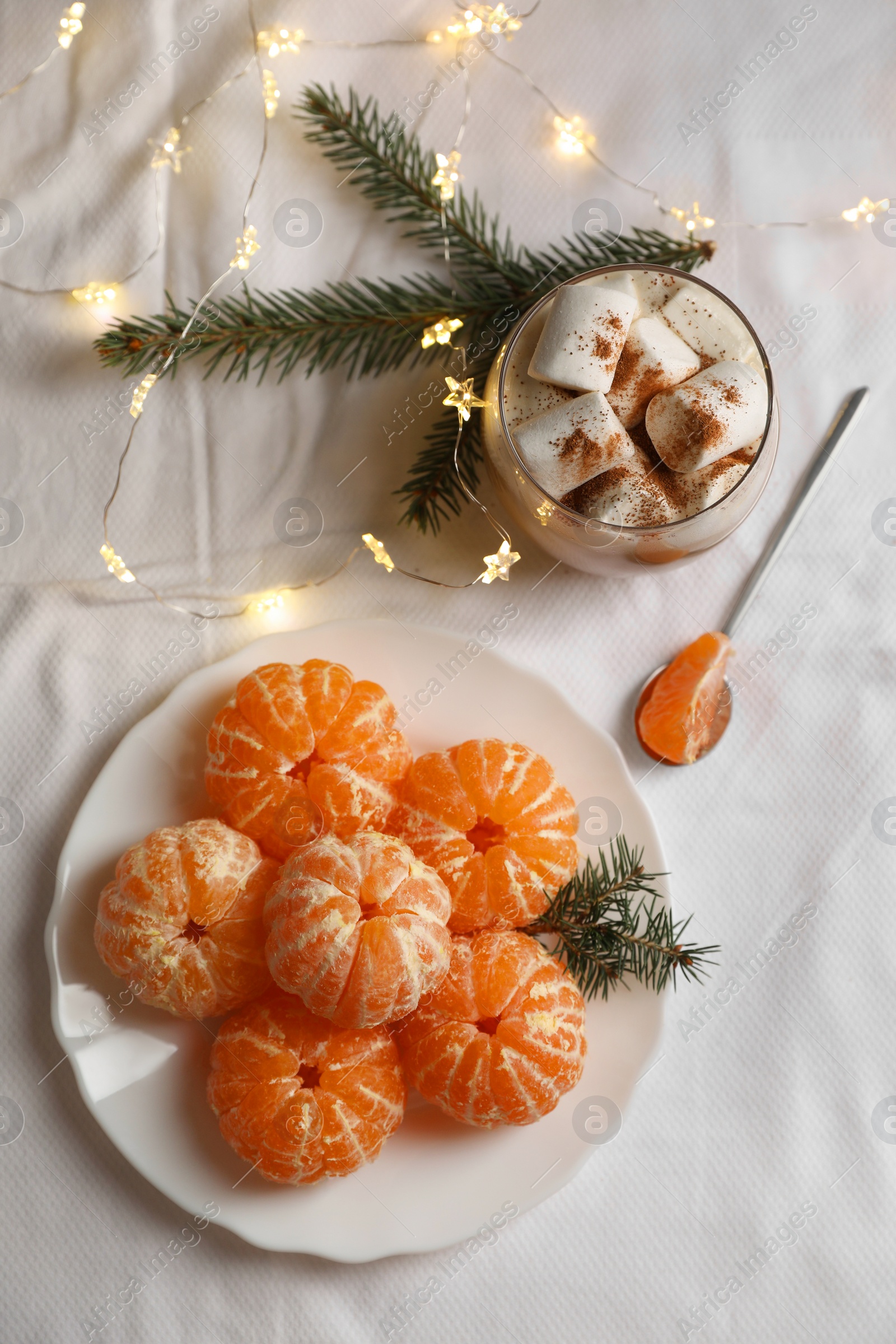 Photo of Peeled delicious ripe tangerines, festive lights and glass of drink with marshmallows on white bedsheet, flat lay