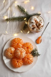 Peeled delicious ripe tangerines, festive lights and glass of drink with marshmallows on white bedsheet, flat lay