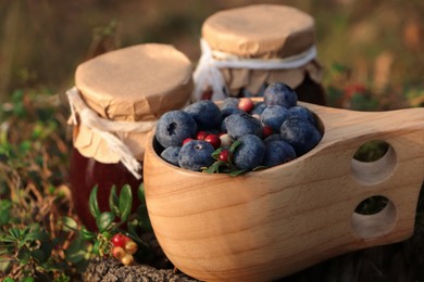Photo of Wooden mug full of fresh ripe blueberries, lingonberries and jars with jam outdoors, closeup